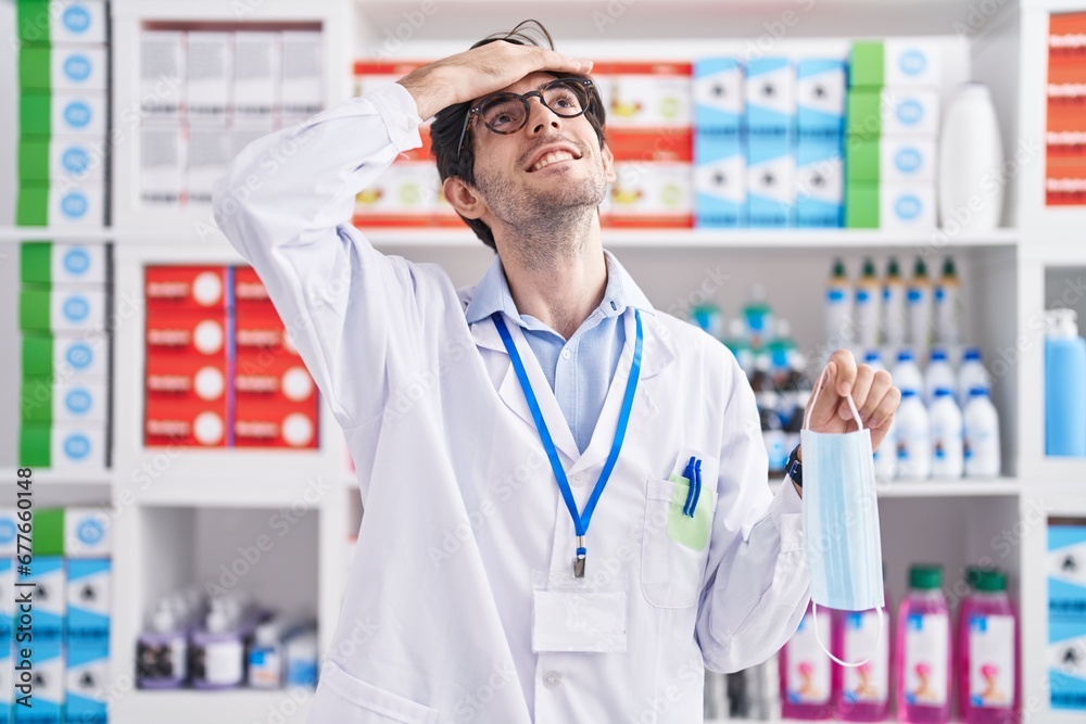 Young hispanic man working at pharmacy drugstore holding safety mask stressed and frustrated with hand on head, surprised and angry face