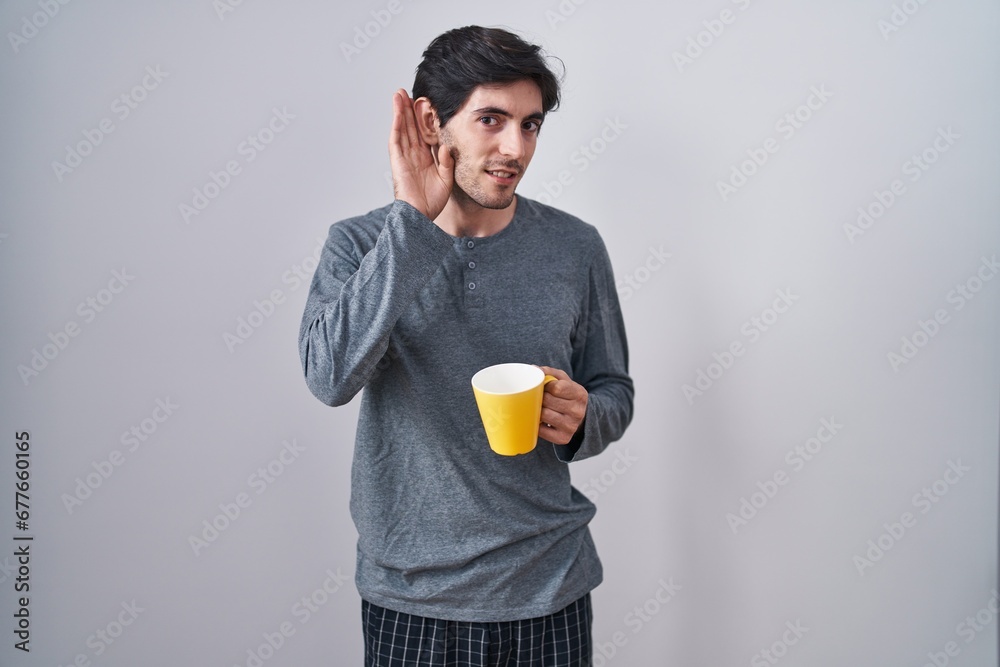 Young hispanic man wearing pajama drinking a cup of coffee smiling with hand over ear listening an hearing to rumor or gossip. deafness concept.