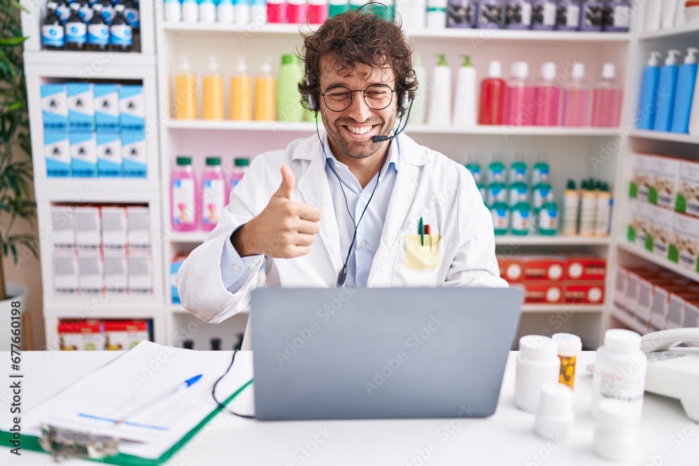 Hispanic young man working at pharmacy drugstore working with laptop smiling happy and positive, thumb up doing excellent and approval sign
