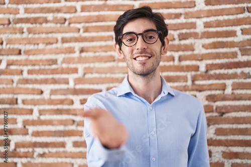 Young hispanic man standing over brick wall background smiling cheerful offering palm hand giving assistance and acceptance.