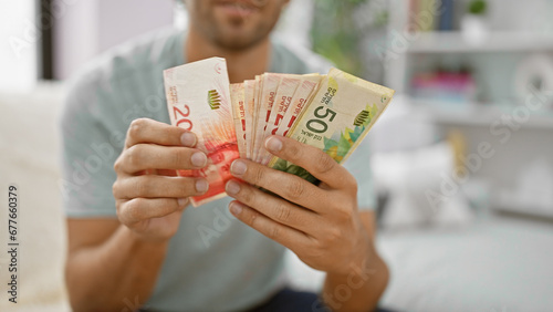 Photos Serious young man immersed in counting stack of israeli shekel banknotes on sofa