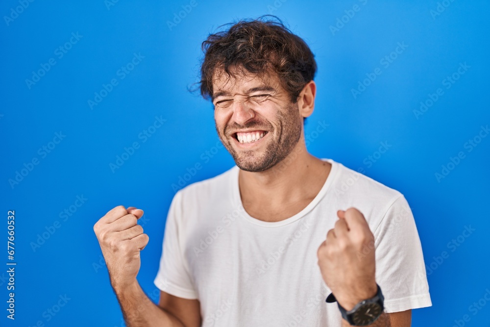 Hispanic young man standing over blue background very happy and excited doing winner gesture with arms raised, smiling and screaming for success. celebration concept. Hispanic young man standing over blue background very happy and excited doing winner gesture with arms raised, smiling and screaming for success. celebration concept.