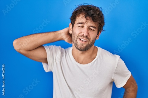 Hispanic young man standing over blue background suffering of neck ache injury, touching neck with hand, muscular pain