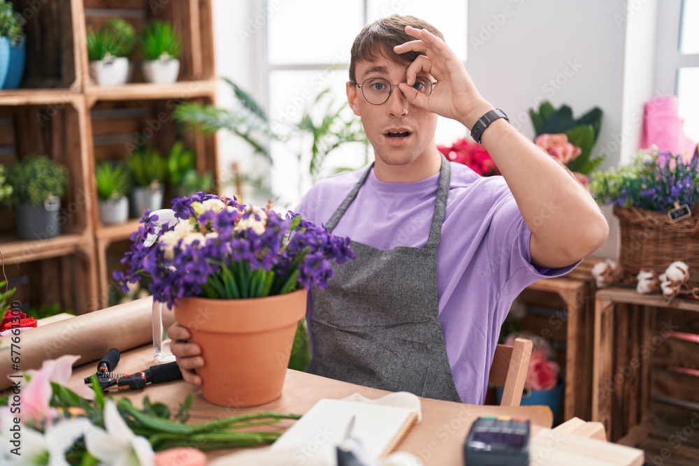 Caucasian blond man working at florist shop doing ok gesture shocked with surprised face, eye looking through fingers. unbelieving expression.