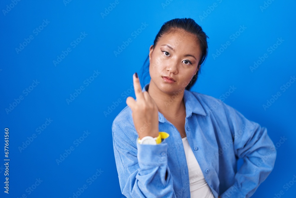 Asian young woman standing over blue background showing middle finger, impolite and rude fuck off expression Asian young woman standing over blue background showing middle finger, impolite and rude fuck off expression