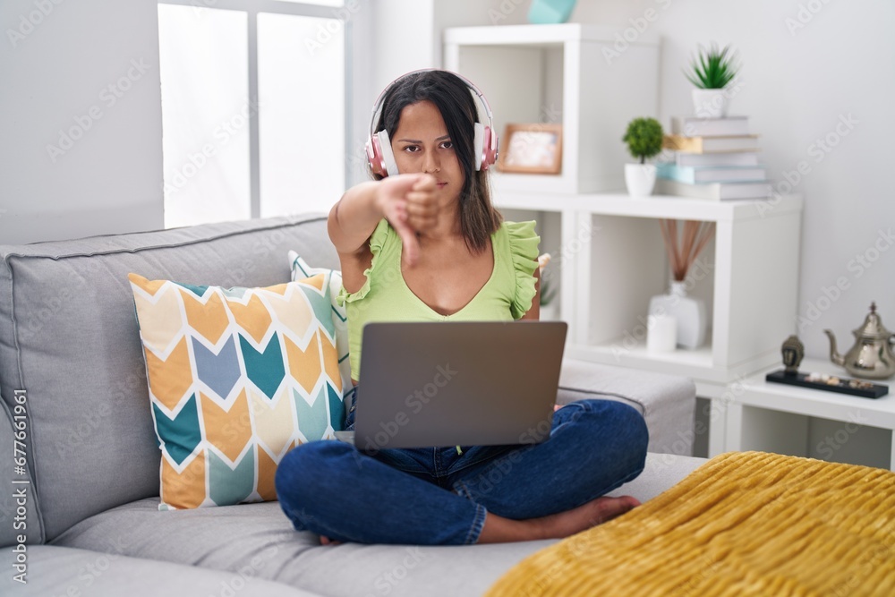 Hispanic young woman using laptop at home looking unhappy and angry showing rejection and negative with thumbs down gesture. bad expression. Hispanic young woman using laptop at home looking unhappy and angry showing rejection and negative with thumbs down gesture. bad expression.