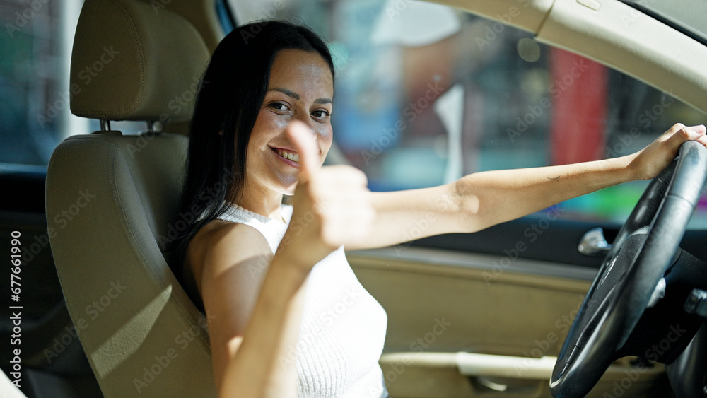 Young beautiful hispanic woman driving car doing ok gesture with thumb up at street