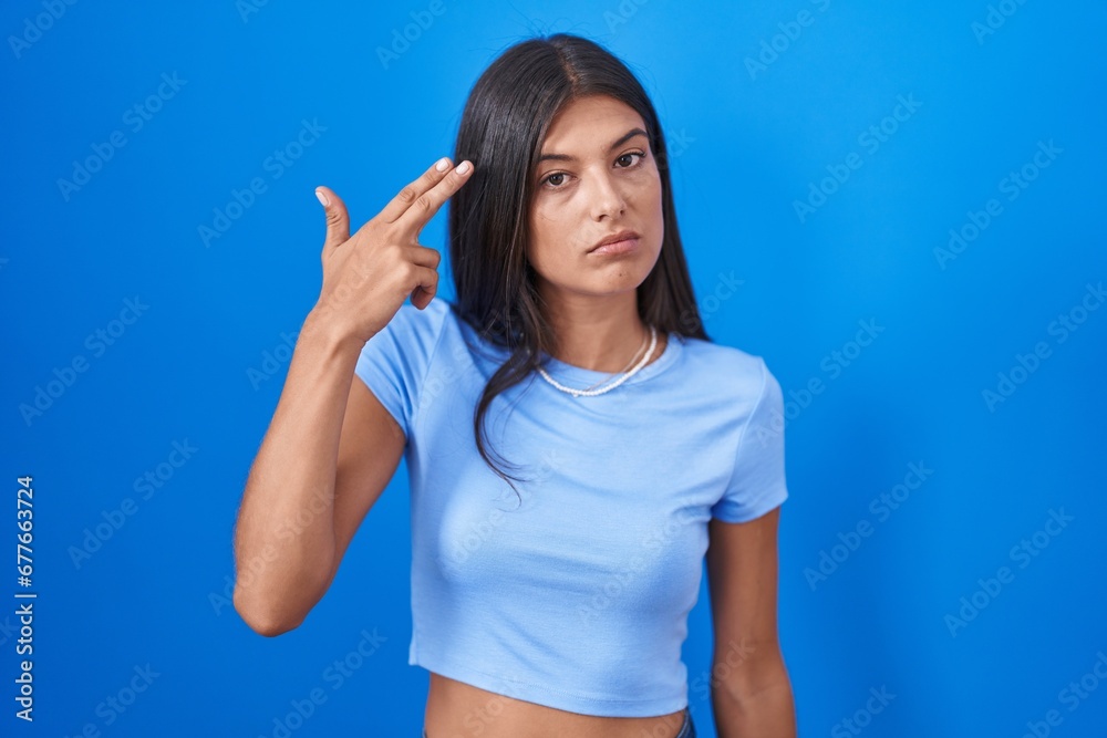 Fototapeta premium Brunette young woman standing over blue background shooting and killing oneself pointing hand and fingers to head like gun, suicide gesture.