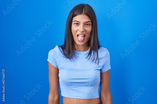 Brunette young woman standing over blue background sticking tongue out happy with funny expression. emotion concept. © Krakenimages.com