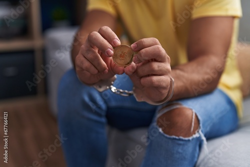 Young hispanic man criminal holding uniswap crypto currency wearing handcuffs at home