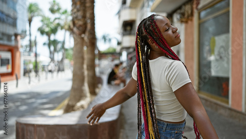Beautiful african american woman with braids, standing on the sunny street, meditating with closed eyes, breathing the summer air, arms open wide, enjoying her freedom, immersed in a relaxed state.