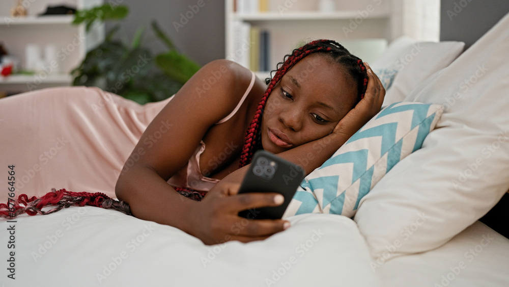 Beautiful african american woman at home, comfortably relaxing on bed in pajamas, typing message using smartphone, amidst a serene bedroom atmosphere