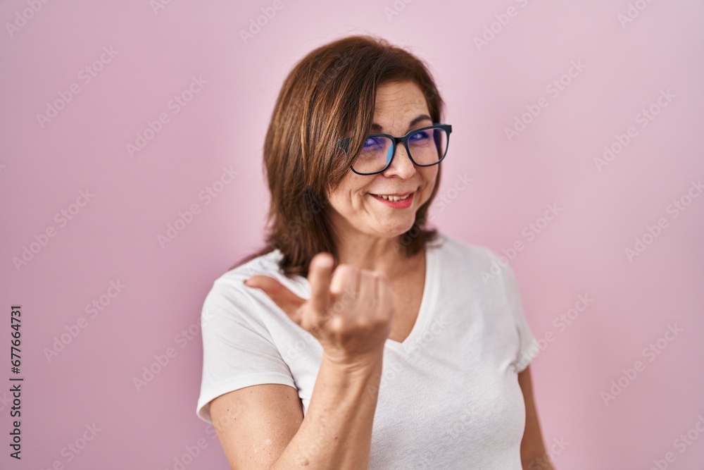 Middle age hispanic woman standing over pink background beckoning come here gesture with hand inviting welcoming happy and smiling Middle age hispanic woman standing over pink background beckoning come here gesture with hand inviting welcoming happy and smiling