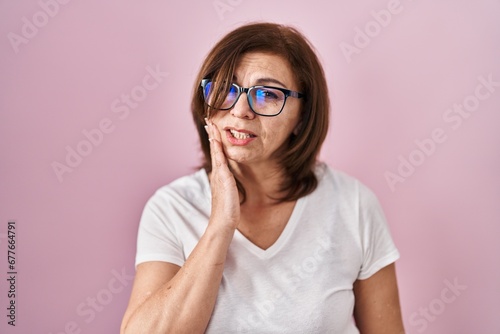 Middle age hispanic woman standing over pink background touching mouth with hand with painful expression because of toothache or dental illness on teeth. dentist