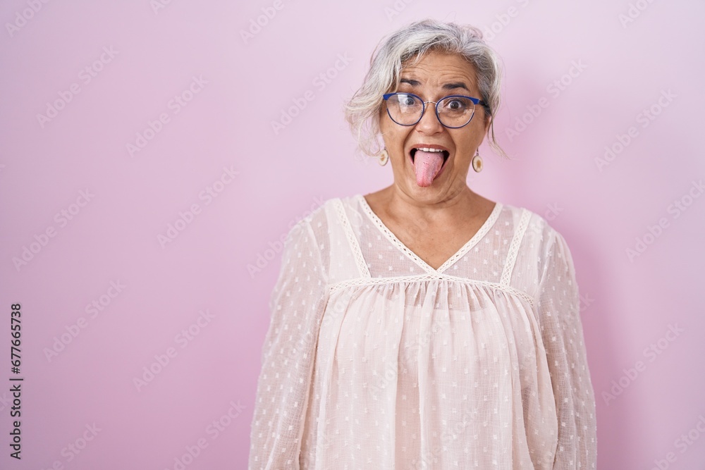 Middle age woman with grey hair standing over pink background sticking tongue out happy with funny expression. emotion concept. Middle age woman with grey hair standing over pink background sticking tongue out happy with funny expression. emotion concept.