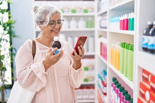 Middle age grey-haired woman customer using smartphone holding medicine bottle at pharmacy photo