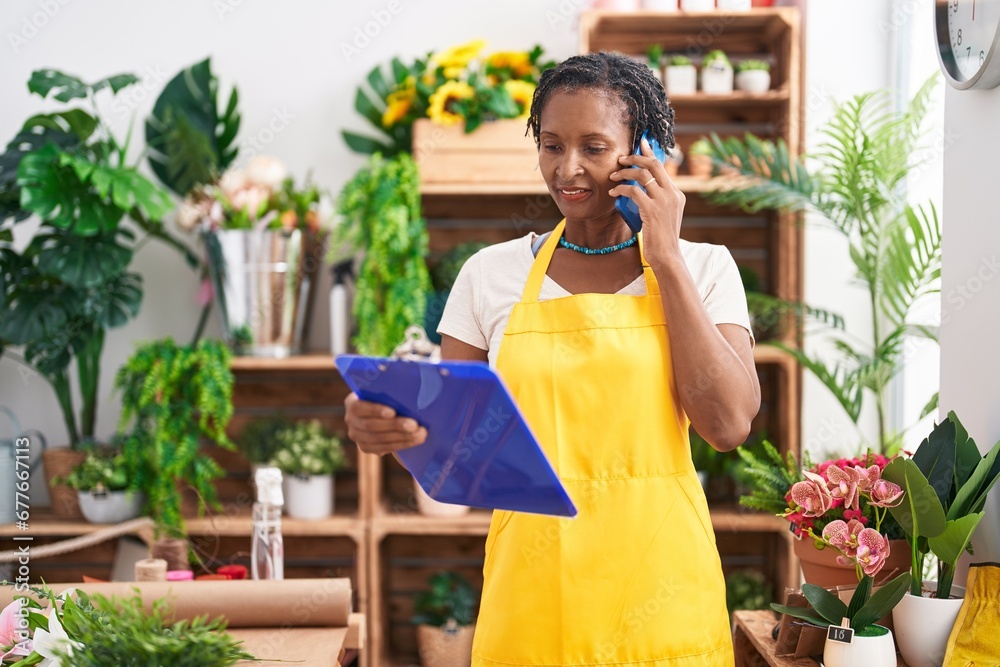 Fototapeta premium Middle age african american woman florist talking on smartphone reading document at flower shop