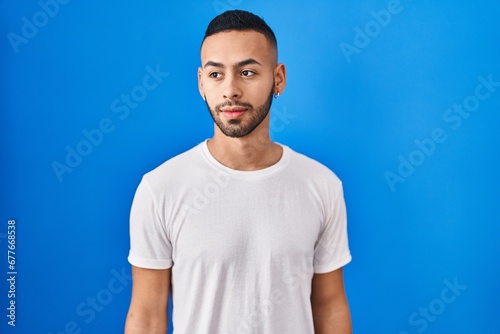 Young hispanic man standing over blue background smiling looking to the side and staring away thinking.