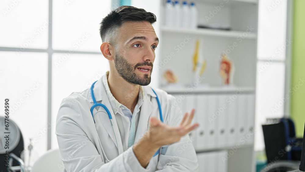 Young hispanic man doctor sitting on table speaking at the clinic Young hispanic man doctor sitting on table speaking at the clinic