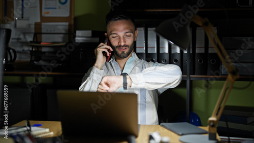 Young hispanic man business worker talking on smartphone looking watch at the office