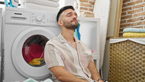 Young hispanic man leaning on washing machine stressed at laundry room
