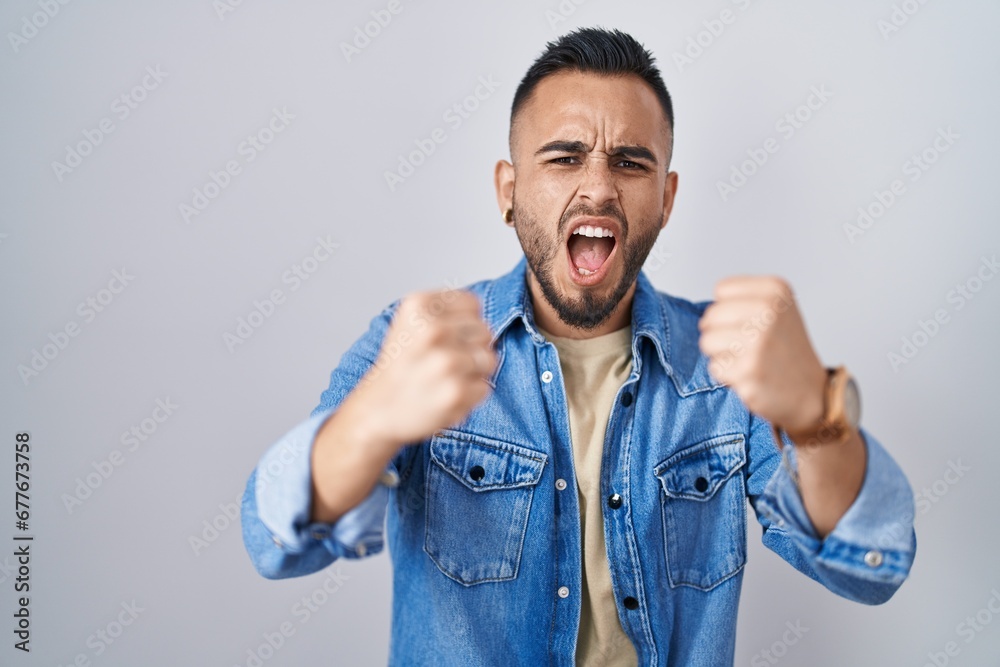 Young hispanic man standing over isolated background angry and mad raising fists frustrated and furious while shouting with anger. rage and aggressive concept. Young hispanic man standing over isolated background angry and mad raising fists frustrated and furious while shouting with anger. rage and aggressive concept.