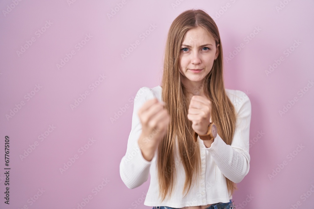 Fototapeta premium Young caucasian woman standing over pink background ready to fight with fist defense gesture, angry and upset face, afraid of problem