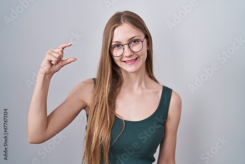 Young caucasian woman standing over white background smiling and confident gesturing with hand doing small size sign with fingers looking and the camera. measure concept.