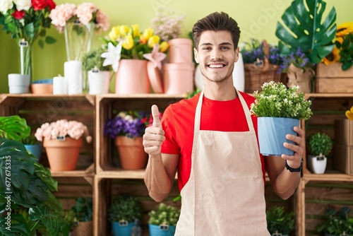 Young hispanic man working at florist shop holding plant smiling happy and positive, thumb up doing excellent and approval sign