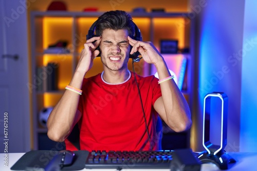 Young hispanic man playing video games with hand on head for pain in head because stress. suffering migraine.