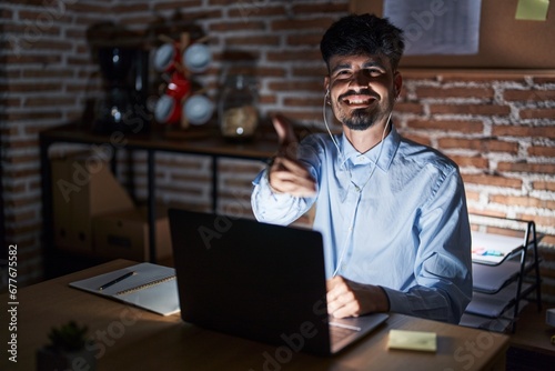 Young hispanic man with beard working at the office at night smiling friendly offering handshake as greeting and welcoming. successful business. photo