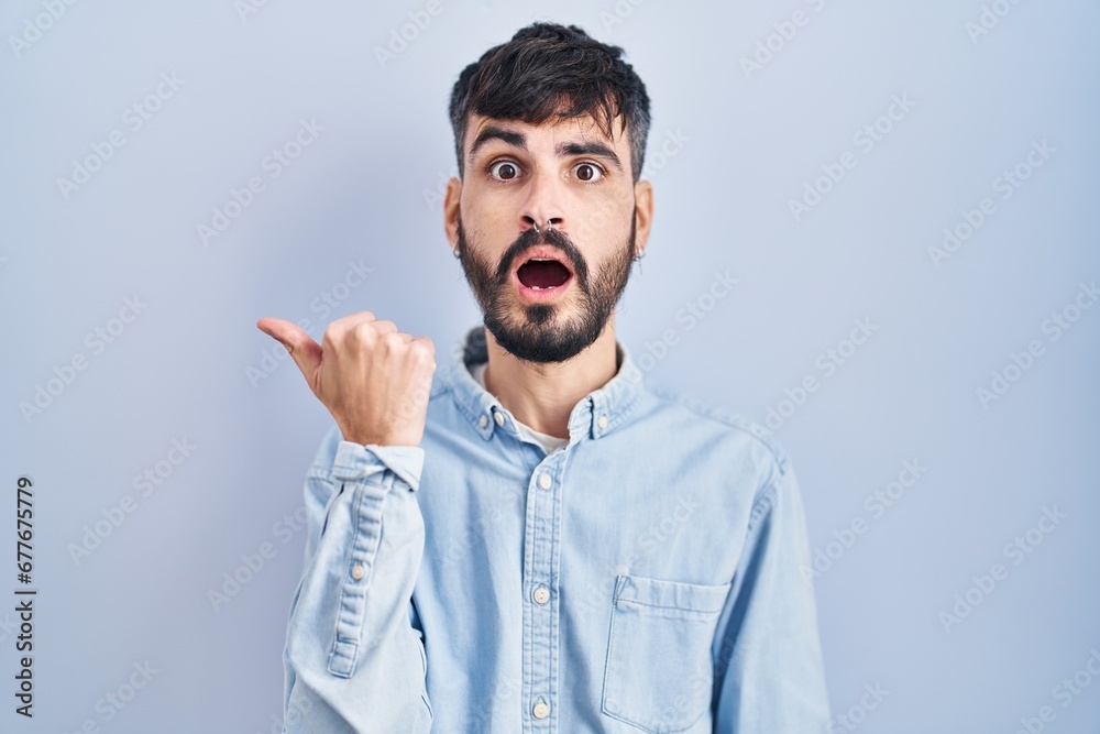 Young hispanic man with beard standing over blue background surprised pointing with hand finger to the side, open mouth amazed expression. Young hispanic man with beard standing over blue background surprised pointing with hand finger to the side, open mouth amazed expression.