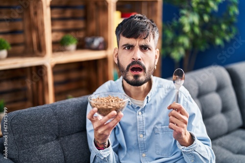 Young hispanic man with beard eating healthy whole grain cereals in shock face, looking skeptical and sarcastic, surprised with open mouth
