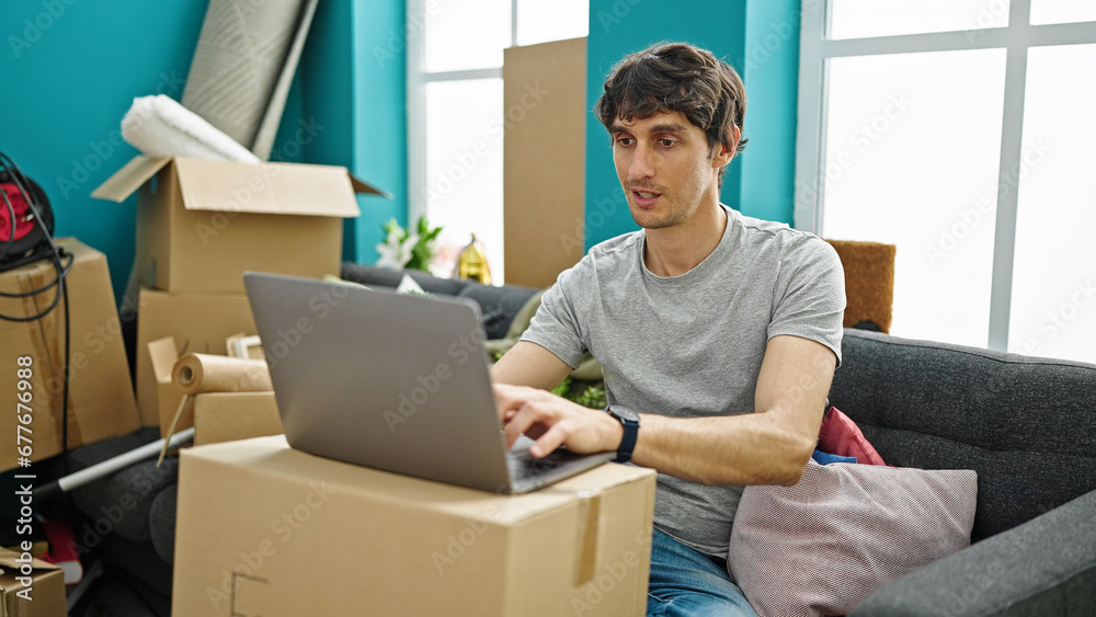 Young hispanic man using laptop sitting on sofa at new home