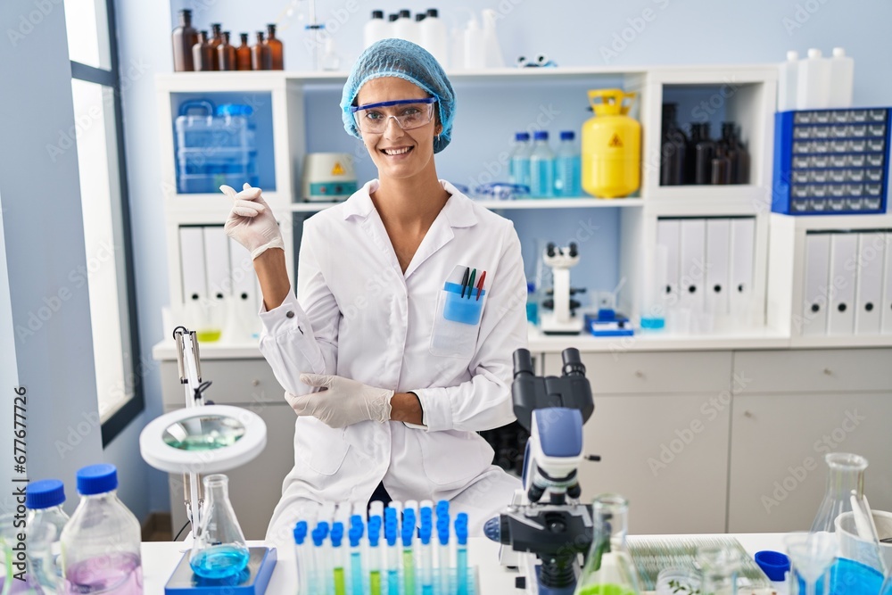 Brunette woman working at scientist laboratory with a big smile on face, pointing with hand finger to the side looking at the camera. Brunette woman working at scientist laboratory with a big smile on face, pointing with hand finger to the side looking at the camera.