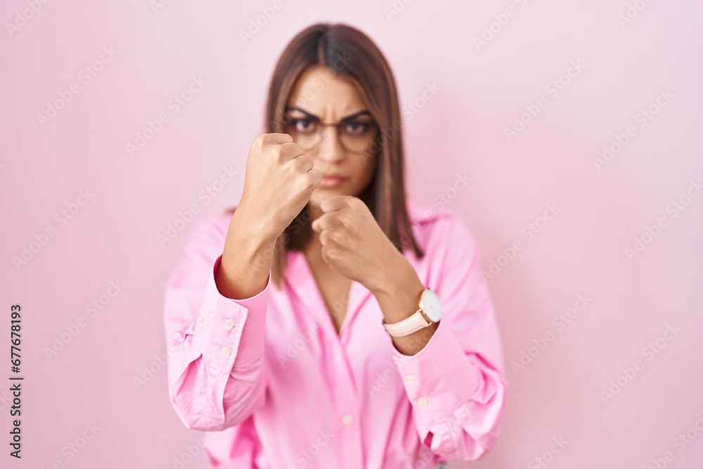 Young hispanic woman wearing glasses standing over pink background punching fist to fight, aggressive and angry attack, threat and violence Young hispanic woman wearing glasses standing over pink background punching fist to fight, aggressive and angry attack, threat and violence