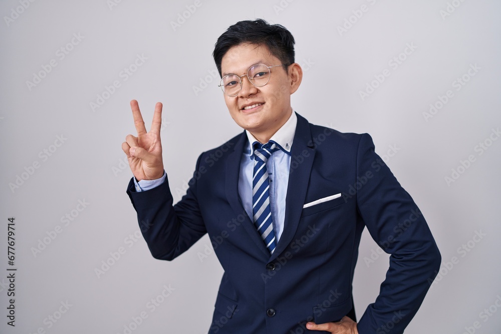 Young asian man wearing business suit and tie smiling looking to the camera showing fingers doing victory sign. number two. Young asian man wearing business suit and tie smiling looking to the camera showing fingers doing victory sign. number two.