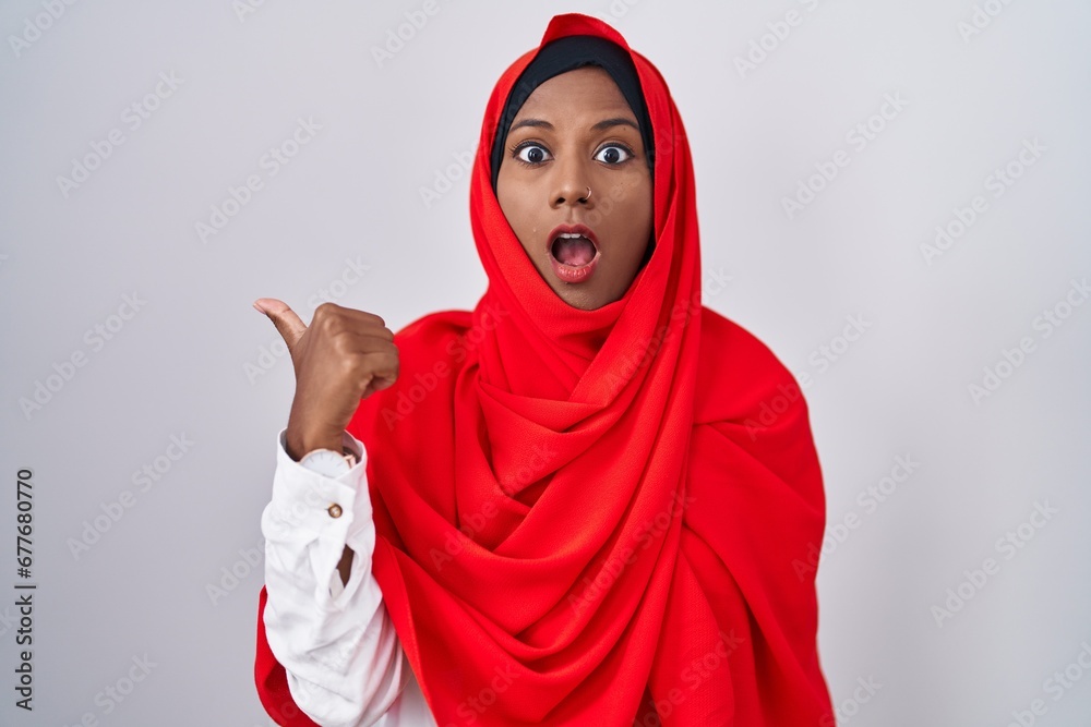 Young arab woman wearing traditional islamic hijab scarf surprised pointing with hand finger to the side, open mouth amazed expression. Young arab woman wearing traditional islamic hijab scarf surprised pointing with hand finger to the side, open mouth amazed expression.