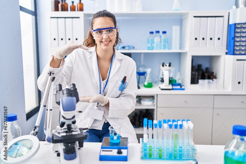Young hispanic woman working at scientist laboratory gesturing with hands showing big and large size sign, measure symbol. smiling looking at the camera. measuring concept. Young hispanic woman working at scientist laboratory gesturing with hands showing big and large size sign, measure symbol. smiling looking at the camera. measuring concept.