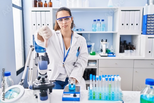 Young hispanic woman working at scientist laboratory looking unhappy and angry showing rejection and negative with thumbs down gesture. bad expression.