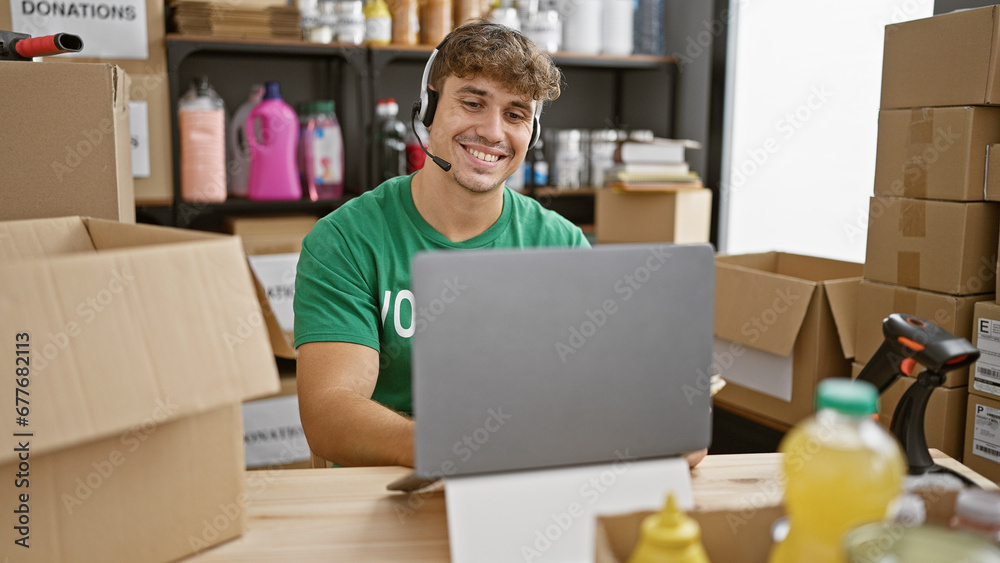 Handsome young hispanic man volunteering at a community charity center, cheerfully smiling while sitting at a table, using laptop with headphones Handsome young hispanic man volunteering at a community charity center, cheerfully smiling while sitting at a table, using laptop with headphones