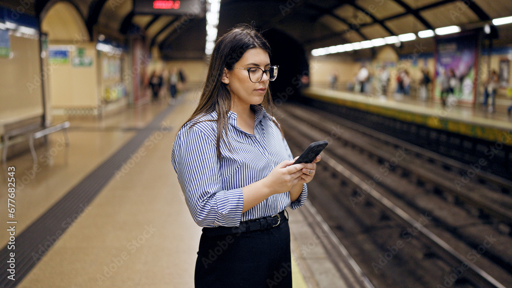 Fototapeta premium Young beautiful hispanic woman waiting for the subway using smartphone in subway station of Madrid