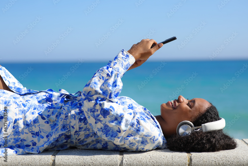 Happy black woman lying on the beach listening audio on phone - obrazy, fototapety, plakaty 