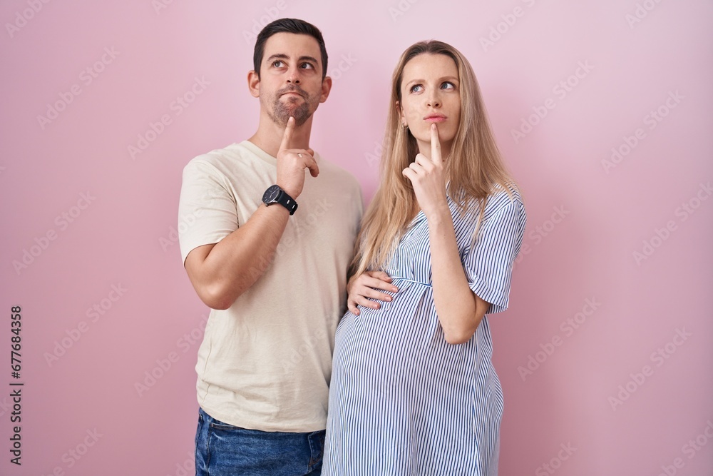 Young couple expecting a baby standing over pink background thinking concentrated about doubt with finger on chin and looking up wondering