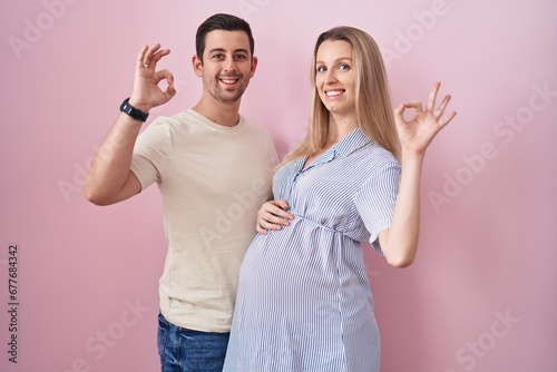 Young couple expecting a baby standing over pink background smiling positive doing ok sign with hand and fingers. successful expression.