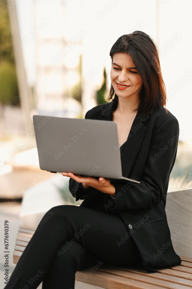 Young business lady working at laptop outdoor on bench near business center. Young business lady working at laptop outdoor on bench near business center.
