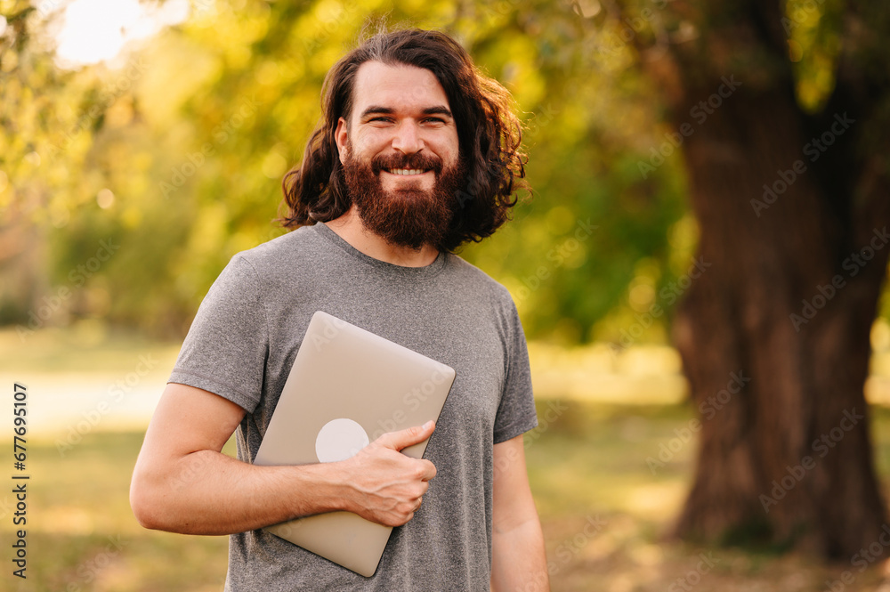 joyful young bearded hipster man holding laptop in park and looking at camera. joyful young bearded hipster man holding laptop in park and looking at camera.
