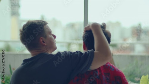 Affectionate gay man stroking hair of beloved boyfriend, then kissing him with love while sitting together by the window at home. View from behind photo