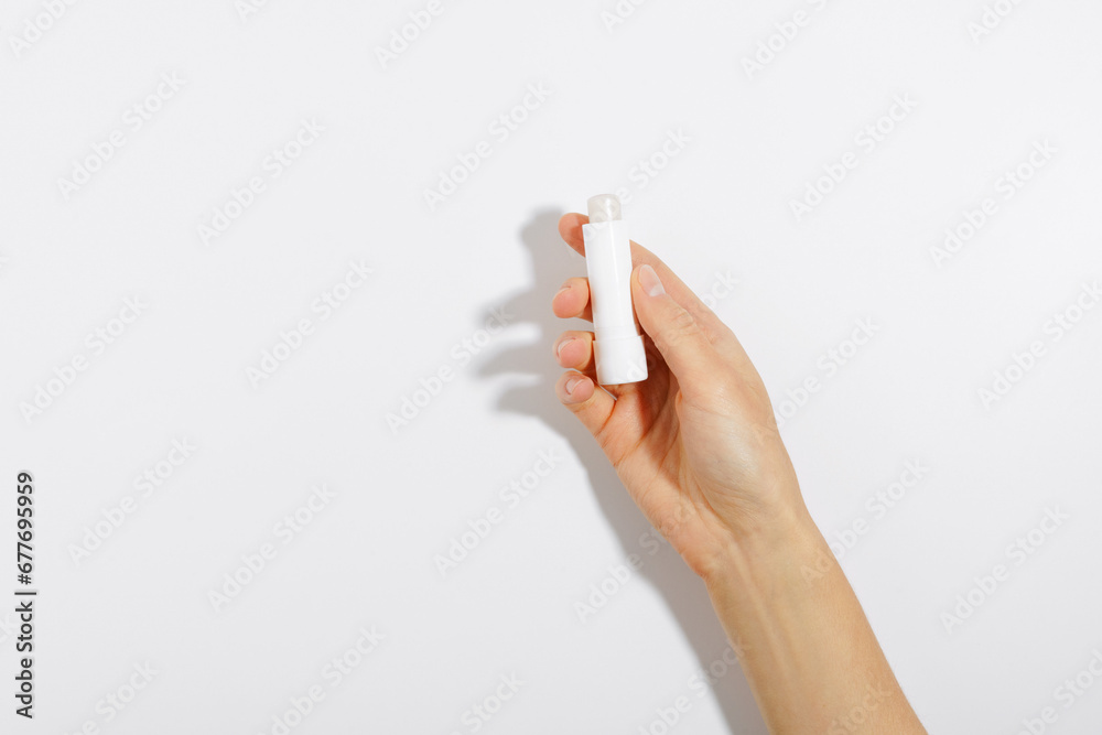 Female hand holding white moisturizing lip balm on white isolated background with shadow from direct light. The concept of beauty products and decorative cosmetics for the face - obrazy, fototapety, plakaty 