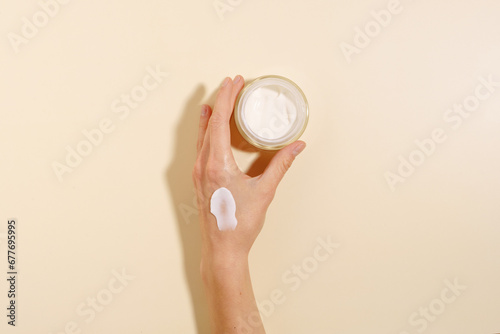 Female hand with cream smear holding glass jar of moisturizer on beige isolated background. The concept of moisturizing and nourishing the skin of the face and body, beauty products
